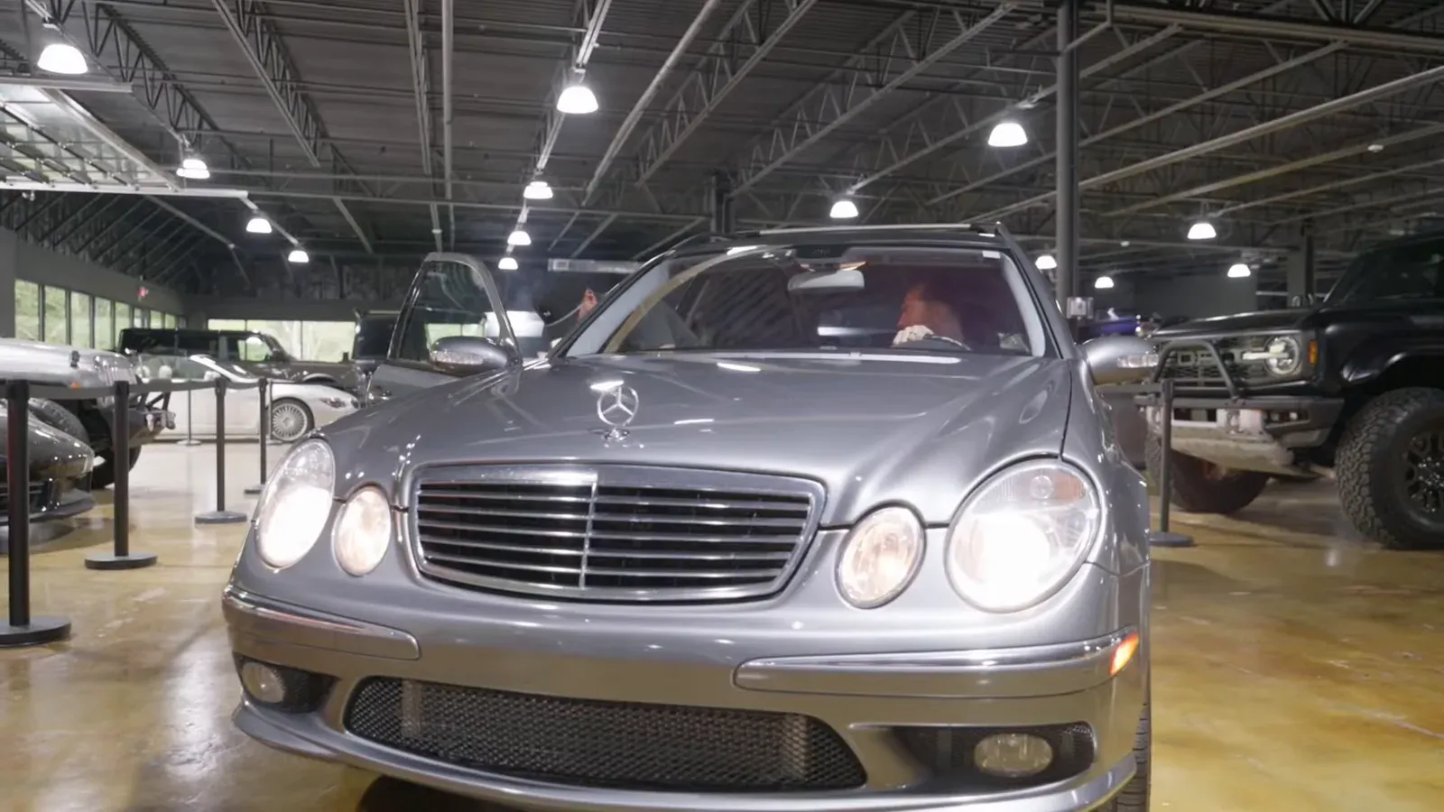 A silver Mercedes E55 AMG wagon parked on the lounge floor with its headlights on, a black truck behind it