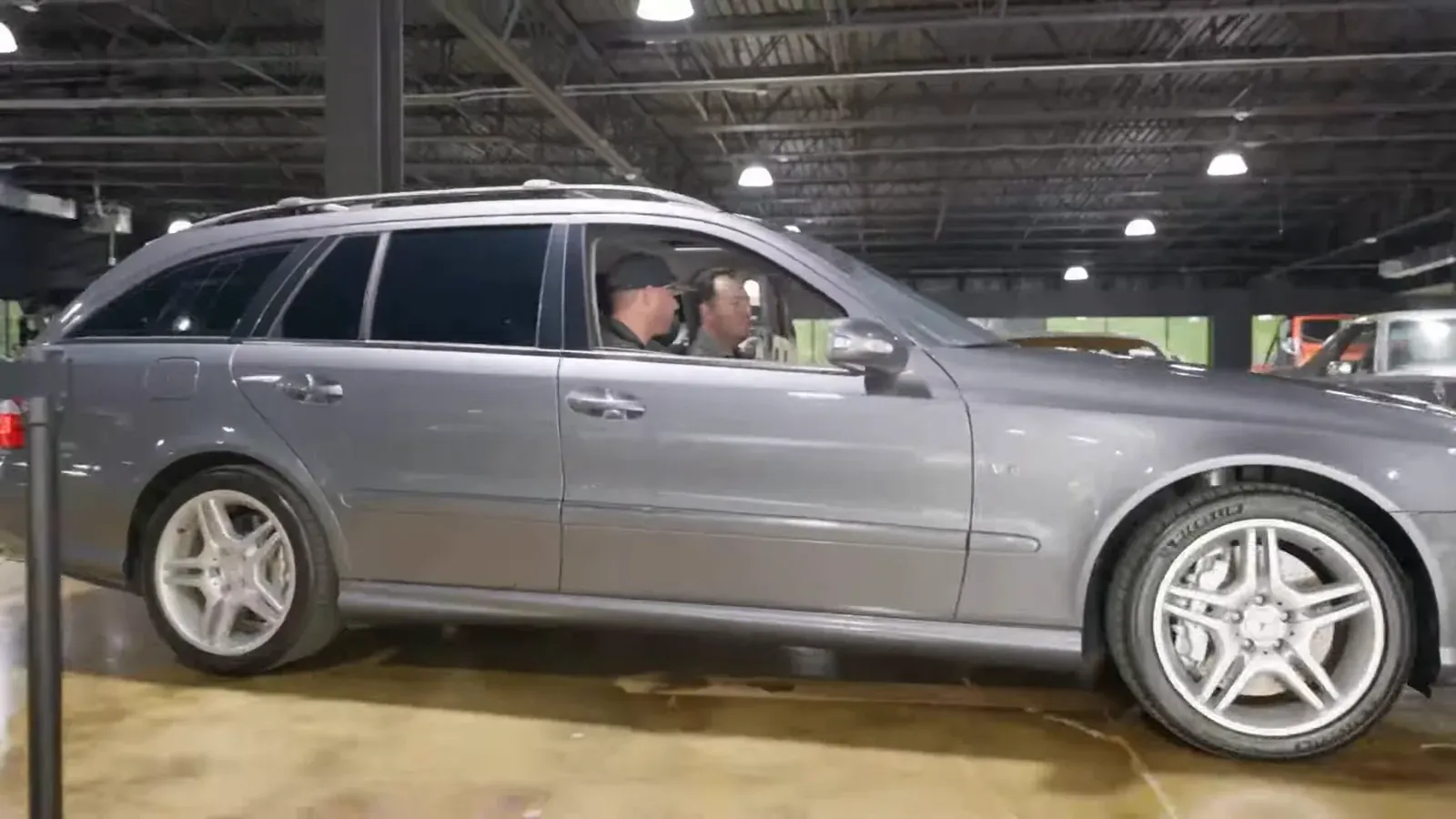 A silver Mercedes E55 AMG wagon rolling across the lounge floor in profile, two men inside