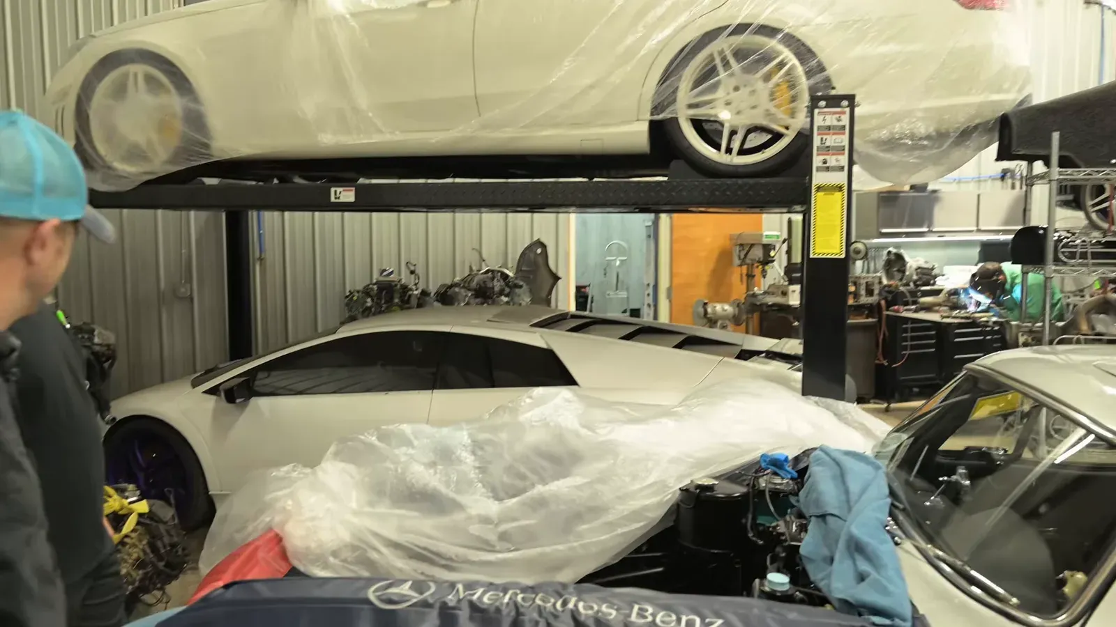 A white Lamborghini Murciélago partly under plastic sheeting on the DC MotorWerks shop floor, a white Mercedes wagon on the lift above it
