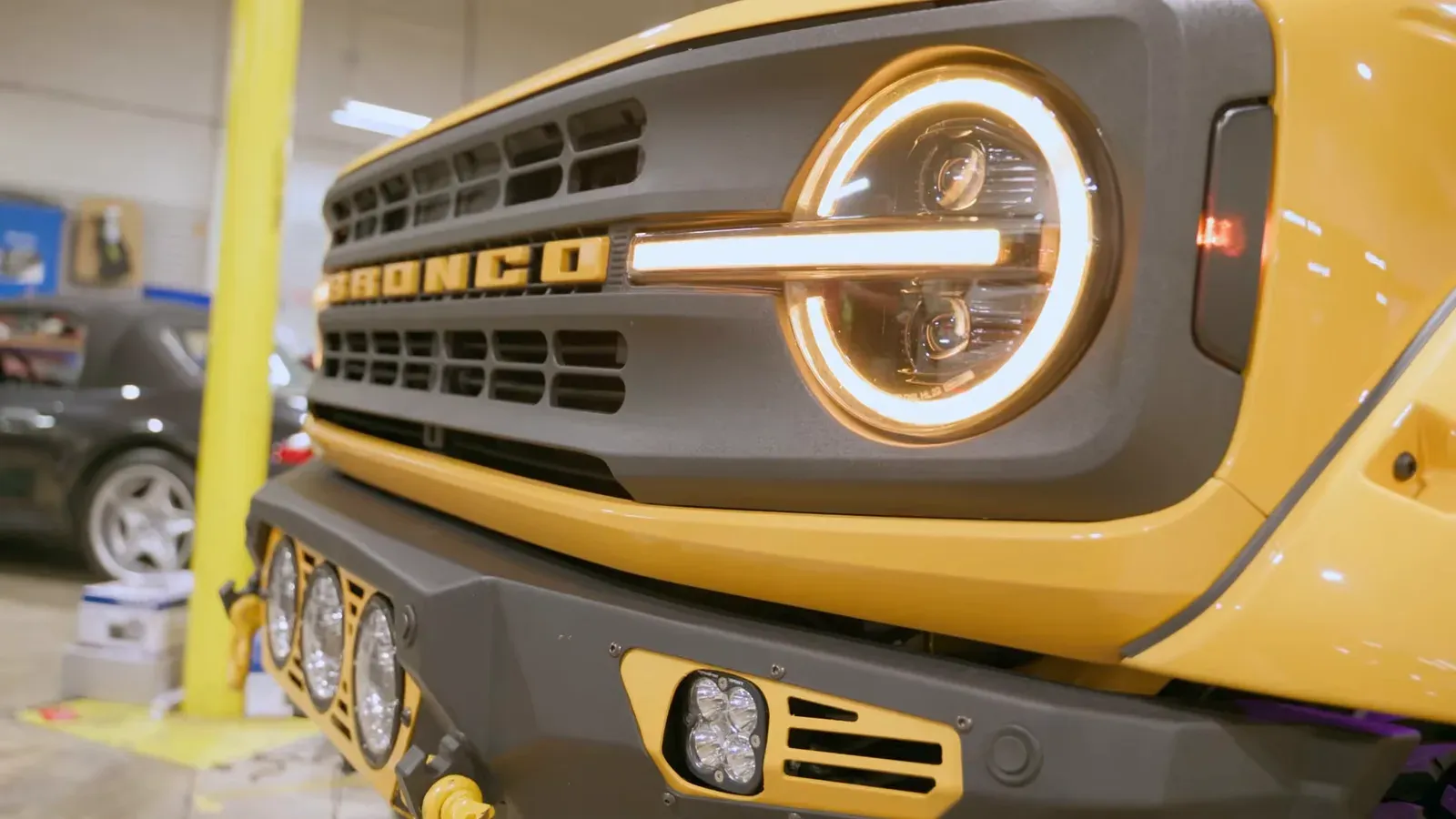 Close-up of the front grille and round LED headlight of a yellow two-door Ford Bronco in the Car Tunes Atlanta shop
