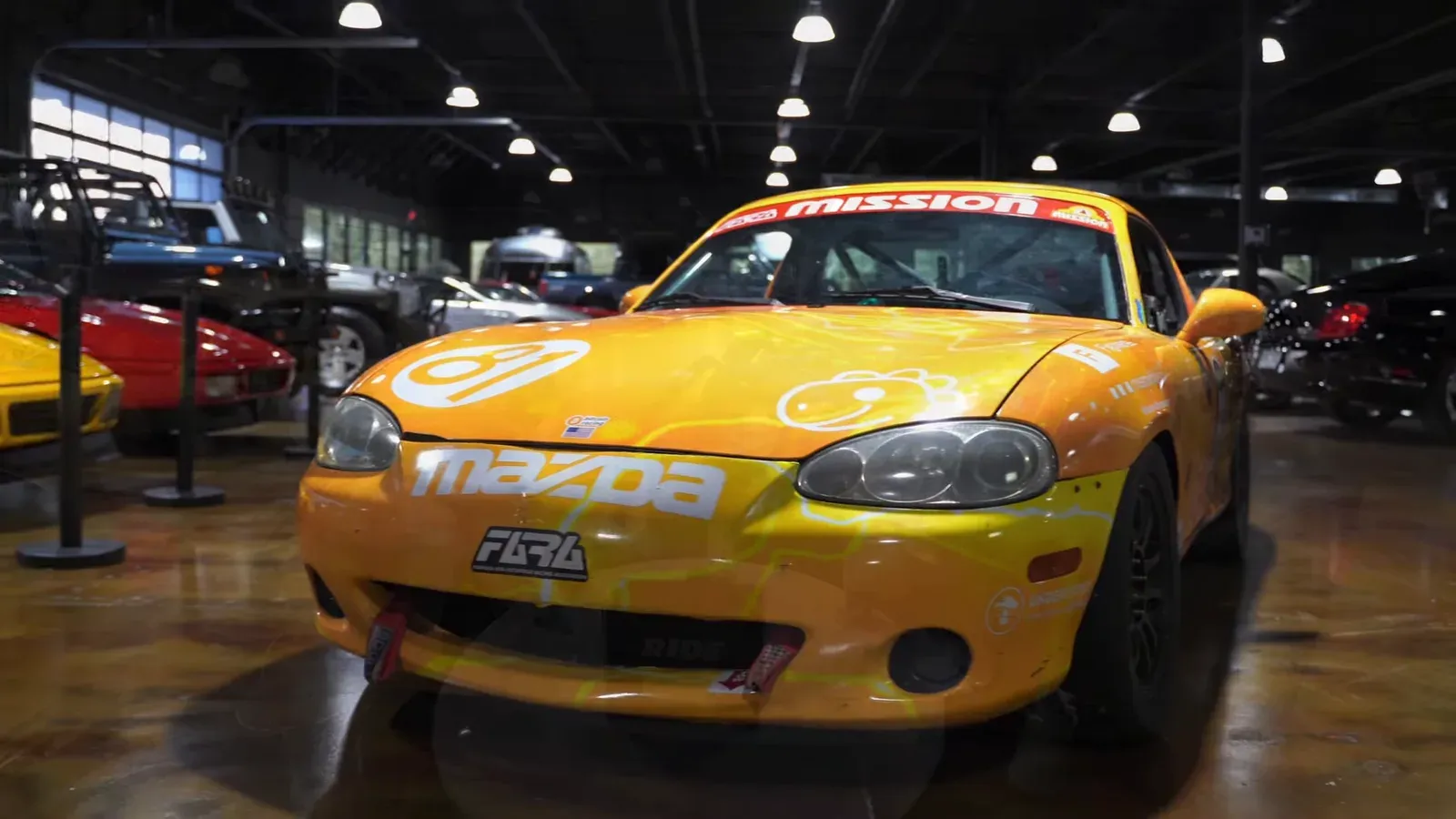 A yellow Mazda Miata race car parked on the lounge floor, seen from the front