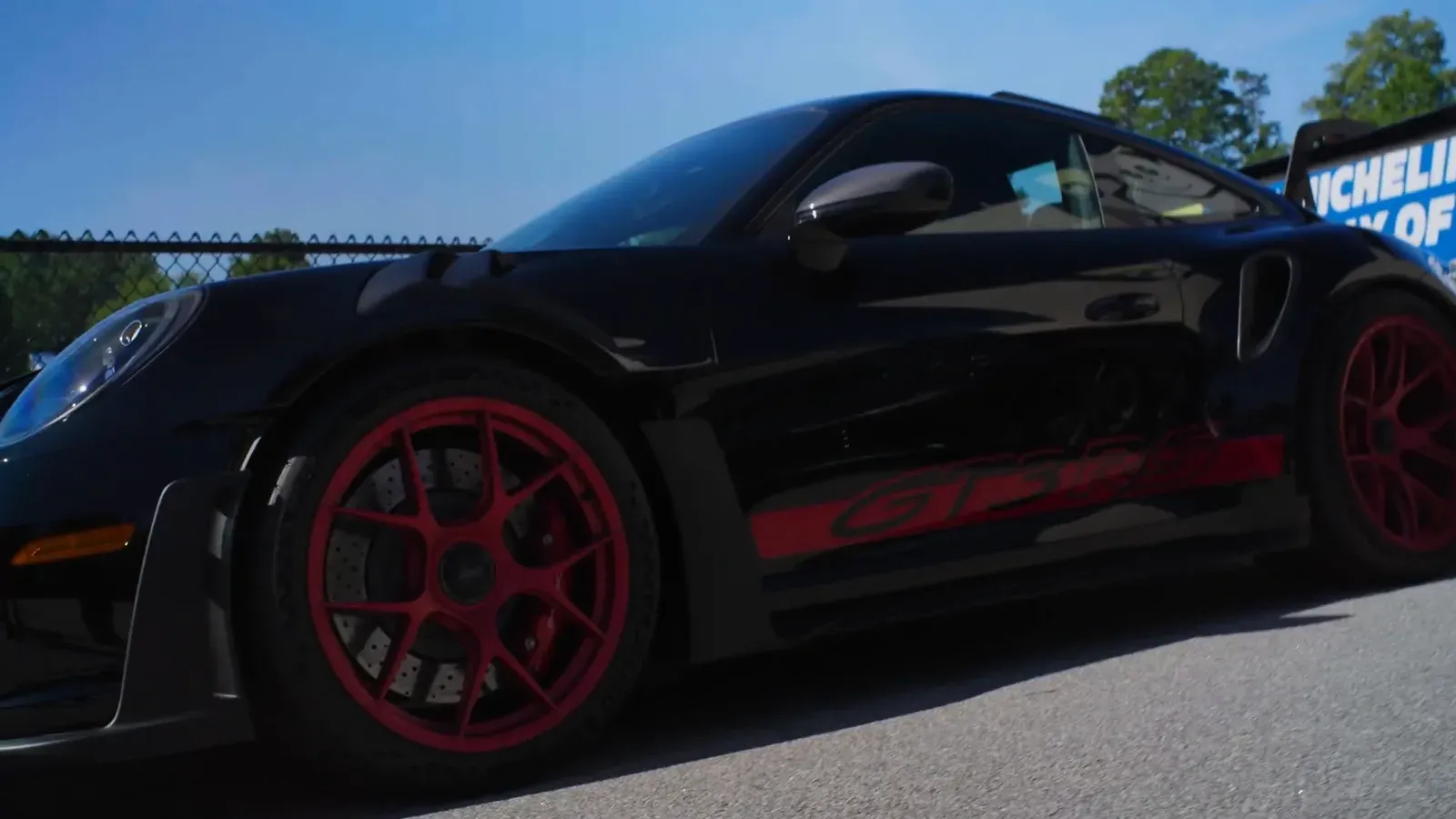 Low side view of a black 2025 Porsche 992 GT3 RS with red wheels and red side stripe, parked at Road Atlanta